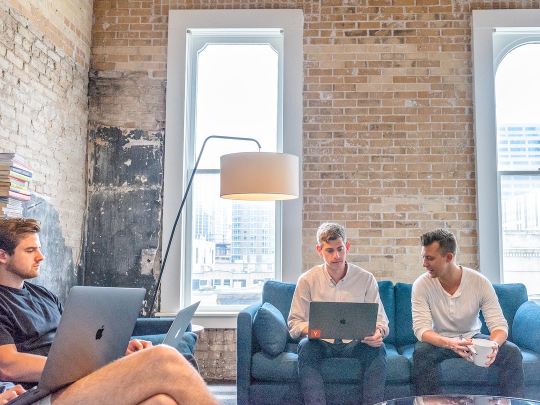 Three men seated on couches, each using a laptop, discussing Knowledge Management in Outsourcing Relationships.